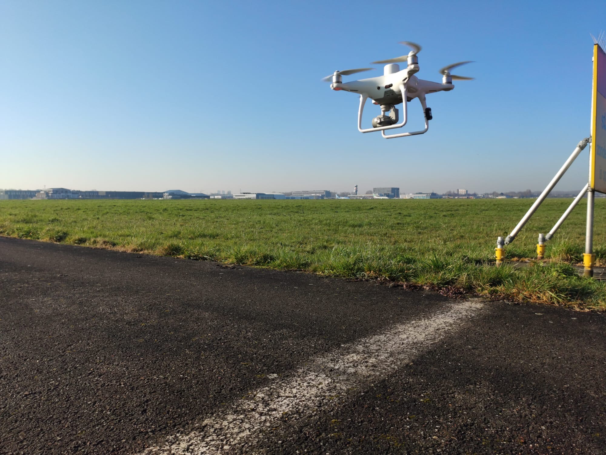 A quadcopter drone hovers over a grassy airfield with buildings in the distance, symbolising unmanned aviation.